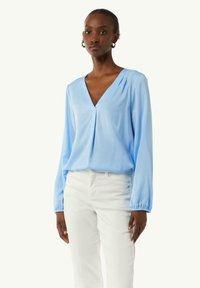 Himmelblause, langärmelige Bluse mit V-Ausschnitt und gerafften Manschetten, aus glattem Stoff gefertigt, locker geschnitten über weißen Hosen.