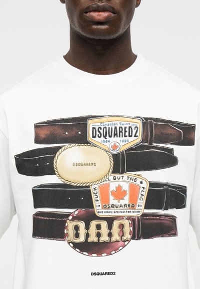 Άνδρας που φοράει λευκό πουκάμισο με εκτύπωση με τέσσερεις οριζόντιες ζώνες με μεγάλες αγκράφες, συμπεριλαμβανομένης της σημαίας του Καναδά και της επωνυμίας DSQUARED2.