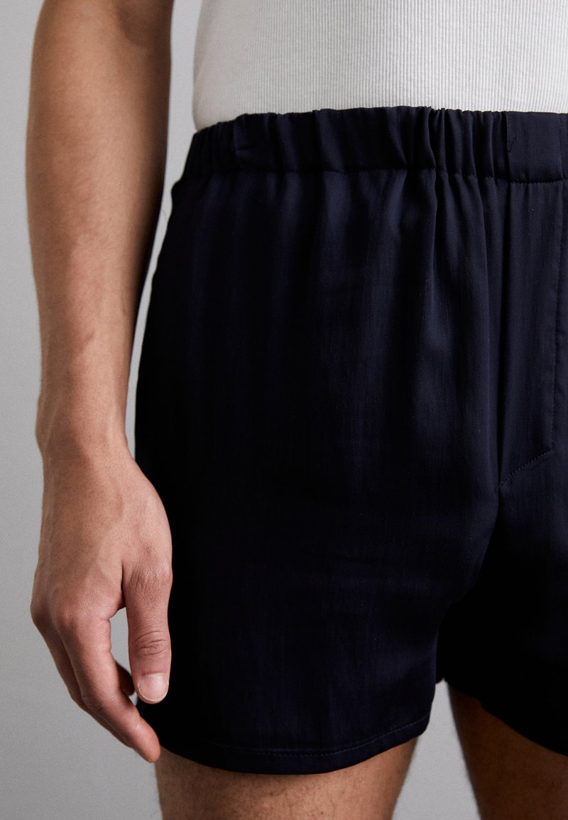 Marineblaue Badeshorts mit einem elastischen Bund, glatter Textur und einer Seitentasche, kombiniert mit einem enganliegenden weißen Tanktop.