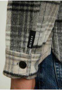 Harmaa ja valkoinen ruudullinen flanelli-paita, jossa on pehmeä rakenne, musta nappisulkeminen ja pieni musta etiketti, jossa lukee "CHASIN'" hihassa.