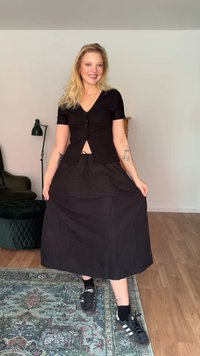 Blonďatá žena se usmívá, drží černou midi sukni, má na sobě černý zapínaný top, tenisky, stojí na vzorovaném koberci v jednoduchém obývacím pokoji.