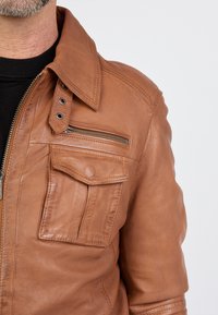 Ruskea nahkatakki, jossa on vetoketjulla suljettava etuosa, suuri rintatasku ja napsautettava kaulus. Sileä pinta, muotoiltu design ja kestävä varustus.