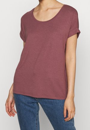 Vrouw met een mauvekleurig t-shirt met korte mouwen, omgeslagen mouwboorden en ronde hals, en een blauwe spijkerbroek tegen een effen achtergrond.
