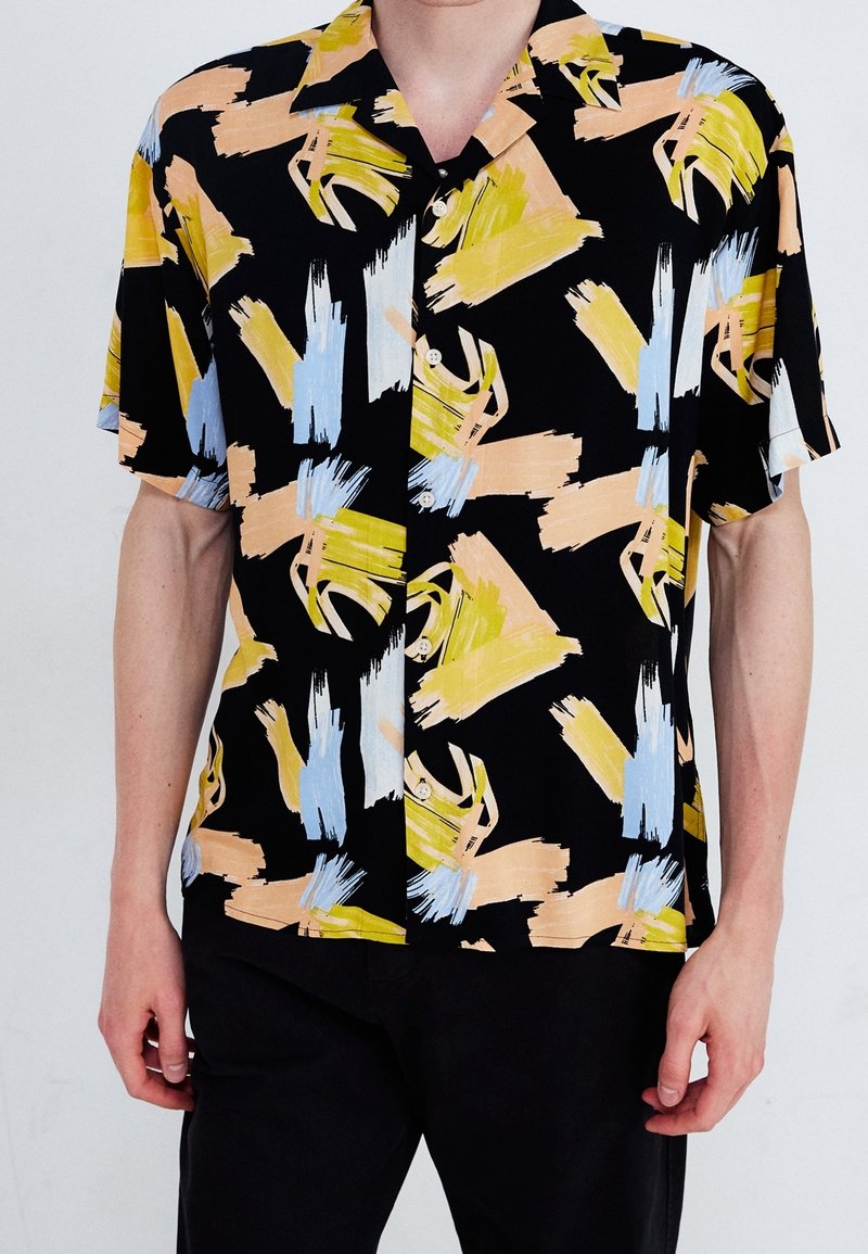 Chemise noire à manches courtes avec des motifs abstraits de coups de pinceau en jaune, bleu et pêche portée par une personne à la peau claire et portant un pantalon noir.