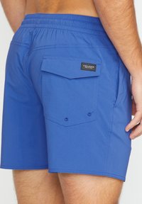 Shorts de bain bleus en tissu lisse, avec un waistband élastique, une poche arrière et des détails de couture renforcée. Logo Volcom attaché.