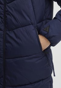 Marineblauer Steppmantel mit einem dicken, gesteppten Design, Reißverschluss auf der Vorderseite und seitlichen Taschen. Textil mit glatter Oberfläche. Akzentuiert mit einem Logopatch.