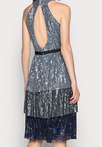 Vestito a balze scintillante con collo all'americana, caratterizzato da un'apertura a goccia sulla schiena e una cintura nera in vita. Paillettes in varie tonalità di blu e argento su tutta la superficie.