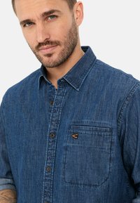 Mørkeblå denimskjorte med knaplukning foran, brystlomme og brune detaljer på lommen. Har en klassisk krave og oprullede ærmer.
