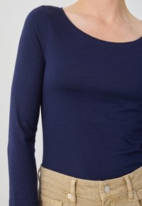 Dugi rukav tamnoplava majica od mekog materijala, sa izrezom u obliku uvala, u kombinaciji sa svijetlosmešanim hlačama i vidljivim šavnim detaljima.