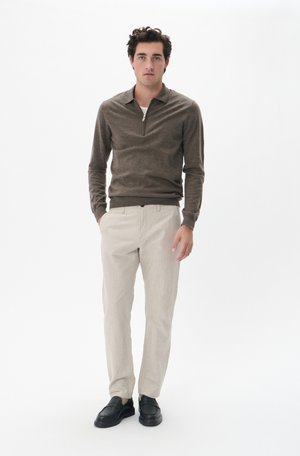 Brun zip-neck sweater med ribbede manchetter, parret med lyse beige bukser. Modellen har sorte loafers på og står foran en hvid baggrund.