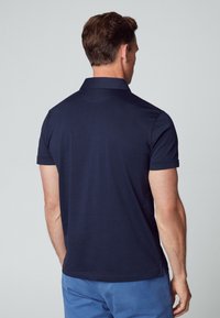 Polo bleu marine en tissu lisse ; doté d'un col classique et de manches courtes, avec des fentes latérales pour faciliter les mouvements.