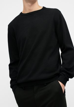 Personne portant un pull noir uni à manches longues avec des poignets côtelés, les mains dans les poches, sur un fond clair uni.