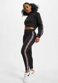 Svart kort hoodie kombinert med svarte leggings med sidestriper i rødt og hvitt tekst. Modellen har på seg hvite Nike-sneakers. Glatt tekstur på stoffet.
