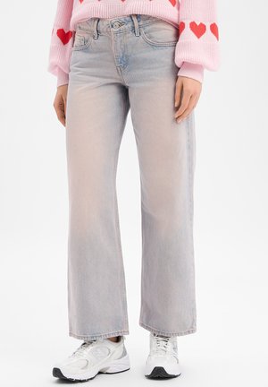 Person trägt hellblaue Jeans mit weitem Bein, weiße Turnschuhe und einen rosa Pullover mit roten Herzen an den Ärmeln.