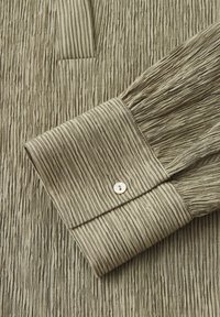 Poignet de chemise texturée à manches longues vert olive clair avec des plis verticaux et un petit bouton blanc sur un fond en tissu assorti.