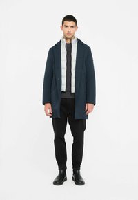 Cappotto lungo blu navy sopra un gilet imbottito grigio, indossato con un maglione lavorato a maglia scuro e pantaloni neri. Presenta bottoni a pressione e un design elegante e minimale.