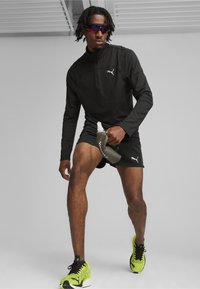 Schwarzes, langärmliges Sportoberteil mit Reißverschluss und Stehkragen, kombiniert mit schwarzen Shorts. Das Model hält eine graue Sportflasche und trägt grüne Turnschuhe.