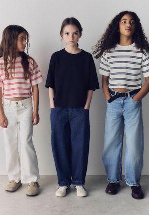 Three girls standing against a white wall, wearing striped cropped tops with jeans and casual shoes, hands in pockets or by sides.