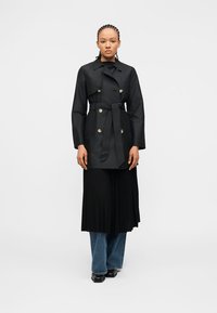 Γυναίκα με πλεξουδωτά μαλλιά που φοράει μαύρο τρανς παλτό με ζώνη πάνω από μαύρη πλισέ φούστα και μπλε τζιν, στέκεται μπροστά σε λευκό απλό φόντο.