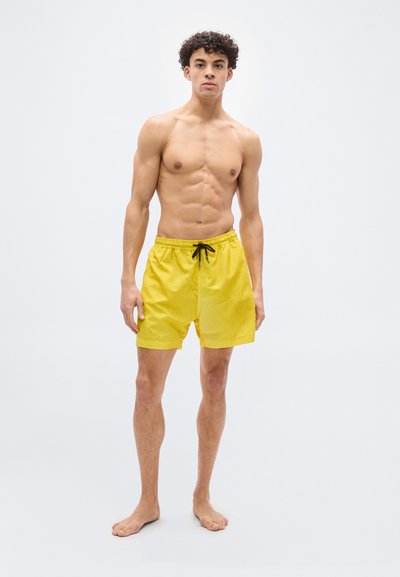 Νεαρός άνδρας στέκεται ξυπόλυτος μπροστά σε λευκό φόντο, φορώντας κίτρινα μαγιό με μαύρο κορδόνι, κοιτάζοντας μπροστά.