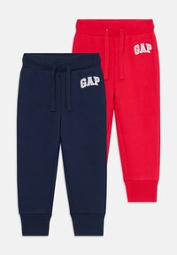 Due pantaloni da bambino piegati, uno di colore blu navy e l'altro rosso, entrambi con vita elastica, cordini e logo bianco "GAP" sull'anca.