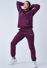 Sudadera con capucha morada con un bolsillo frontal y pantalones de chándal a juego que cuentan con puños elásticos. Zapatillas blancas completan el atuendo.