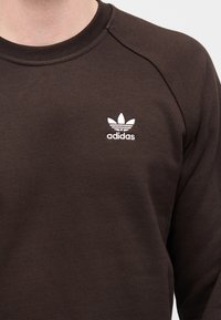 Ruskea collegepaita, joka on valmistettu teksturoidusta kankaasta. Vasemman rinnan alueella on valkoinen Adidas-logo ja kolme lehtistä embleemiä.