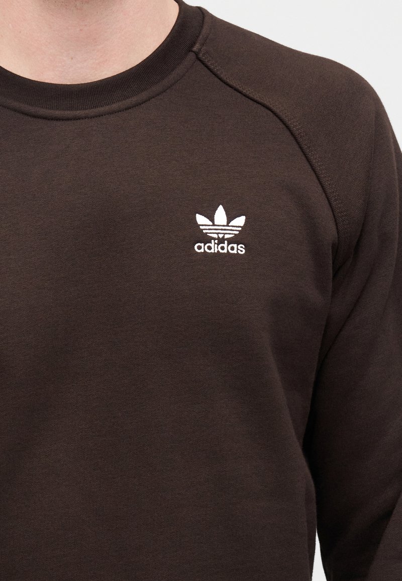 Brun sweatshirt i tyg med texturerad yta, med en vit Adidas-logotyp och trebladig emblem på vänster bröst.