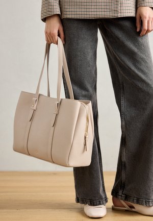 Bolsa tote em pele bege com duas alças superiores, design estruturado, detalhe de fecho dourado e uma marcação subtil, combinada com jeans cinzentos de perna larga.