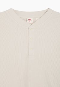 Beige ribbet henley-skjorte med rund krage, tre knapper, og et merke som indikerer "Levi's, Standard Fit." Teksturert stoff.