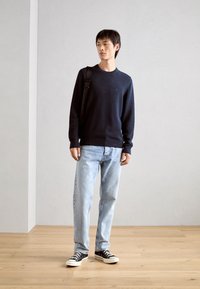 Marineblaues Rundhalspullover, hellblaue Jeans, schwarze Sneaker, stehend mit einer Schultertasche, auf einem Holzboden vor einem neutralen Hintergrund.
