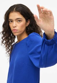 Blusa blu strutturata con maniche a sbuffo e polsini arricciati. Tessuto liscio e leggero. Design minimalista con scollatura rotonda.