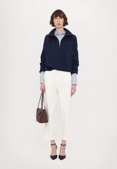Navy pullover with a high collar and silver zip, layered over a light blue striped shirt. White trousers and a brown handbag complete the look.