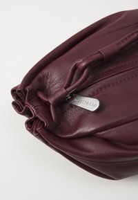 Borsa in pelle color borgogna con design arricciato, sormontata da una maniglia ad anello e un'etichetta accentuata in argento con marchio. Texture liscia con cuciture in contrasto.