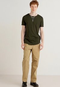 Tricou verde măslin asortat cu chino-uri bej. Hainele au o croială lejeră, o textură netedă și pantofi din piele neagră.