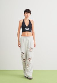 Mustat halter-top crop -paita logolla; harmaat collegehousut, joissa on kuluneet ja repeytyneet yksityiskohdat; malli seisoo vihreällä lattialla yksinkertaista taustaa vasten.
