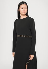 Black long-sleeve dress with gold accents at the waist and side slit. Smooth fabric with a round neckline. Minimalistic design.