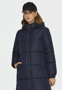 Doudoune bleu marine avec capuche, dotée d'une fermeture éclair intégrale, d'un motif matelassé et de poches latérales ; portée avec une casquette noire.
