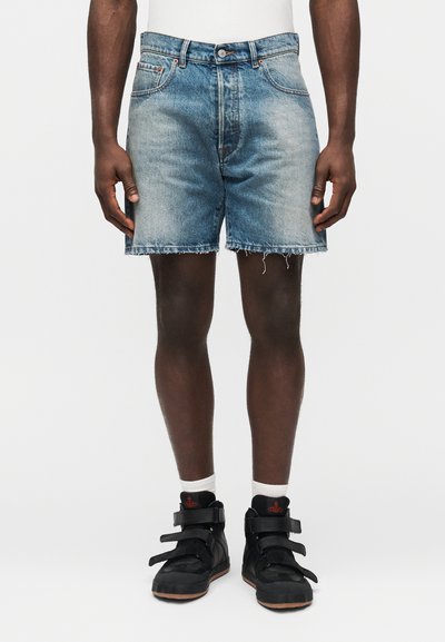 Shorts en jean bleu clair avec un effet délavé, bord effiloché, design classique à cinq poches et fermeture à boutons. Portés avec des baskets noires.