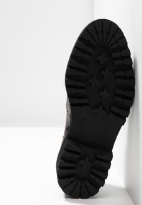 Semelle d'une botte sombre avec des crampons profonds montrant des motifs en forme d'étoile et linéaires, inclinée contre un fond blanc en coin.