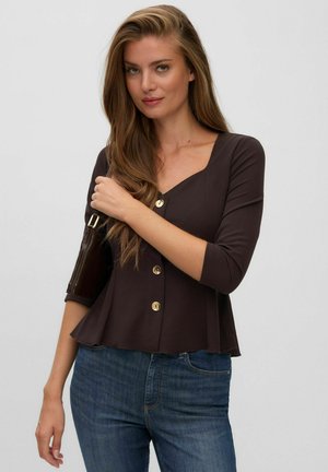 Femme aux longs cheveux châtain clair portant un haut foncé boutonné et un jean bleu, tenant un petit sac à bandoulière marron avec une boucle dorée.