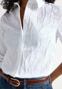 Hvid skjorte med knaplukning og tekstureret, bølget stof. Har en krave og diskrete mønstre. Båret med et brunt bælte og denim jeans.