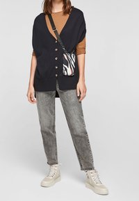 Mörkblå stickad cardigan med knappar, brun skjorta under, grå slitna jeans och mönstrad svart, vit och röd crossbody-väska. Vita sneakers.