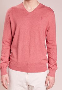 Homme portant un pull corail à col en V et manches longues avec un petit logo brodé sur la poitrine et un pantalon beige clair.