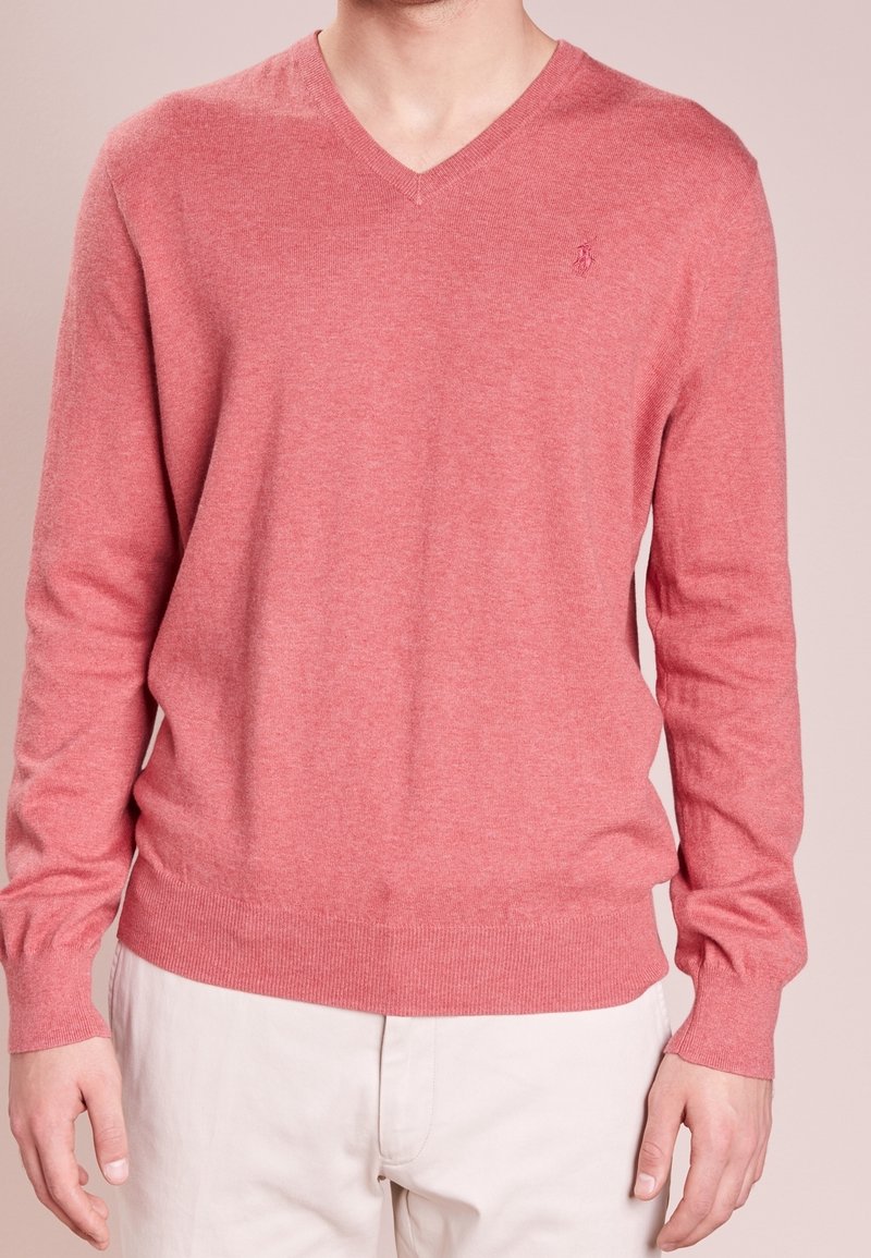 Homme portant un pull corail à col en V et manches longues avec un petit logo brodé sur la poitrine et un pantalon beige clair.