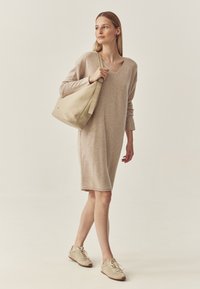 Beige svetr šaty s dlouhými rukávy a výstřihem do V, doplněné béžovými teniskami a velkou, měkkou, světlou kabelkou na rameno.