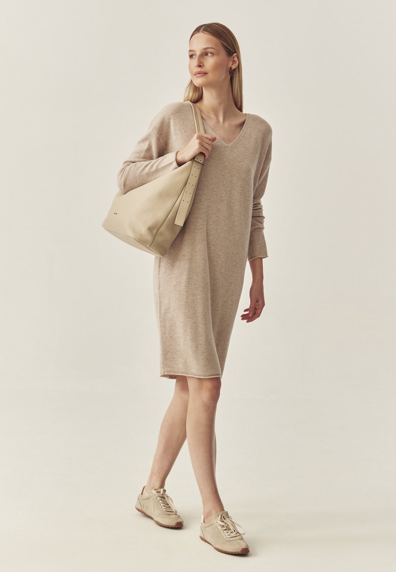 Beżowa sukienka swetrowa z długimi rękawami i dekoltem w kształcie litery V, w zestawie z beżowymi sneakersami i dużą, miękką, jasną torbą na ramię.