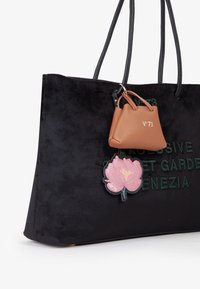 Borsa a spalla in suede nera con testo in rilievo, dotata di un piccolo charm in pelle marrone e di una patch floreale rosa, appesa a una tracolla nera.