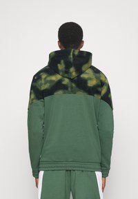 Felpa con cappuccio caratterizzata da un corpo verde con una parte superiore in mimetica nera e verde. La texture è morbida; il design include una cucitura sulle spalle.