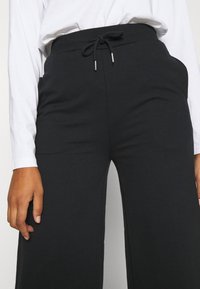 Svarta vida byxor i mjukt material med elastisk midja och dragsko. Fickor syns på sidorna.